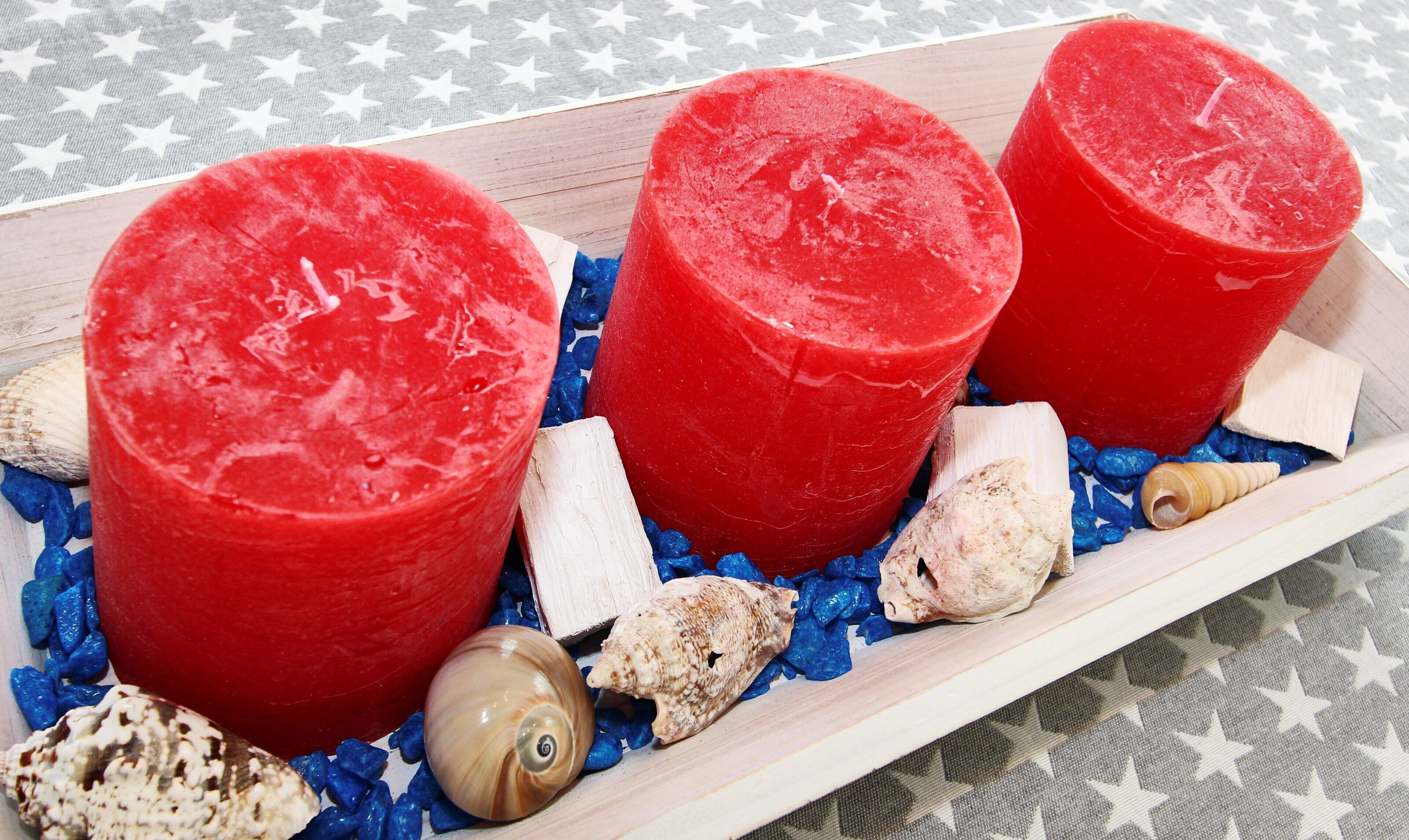 Tres cilindros rojos sobre una bandeja de madera con conchas marinas y guijarros azules.