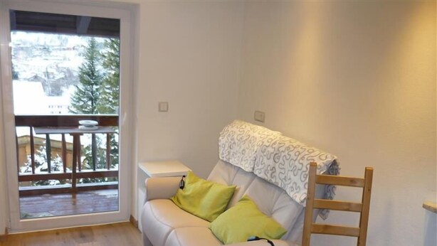 Ein kleiner, gut beleuchteter Raum mit einem weißen Sofa, einem Holzstuhl und einem kleinen Tisch. Durch eine offene Tür ist ein Balkon mit Geländer zu sehen.