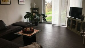 A living room with a dark leather sofa, a small table with a decorative item, a television on a cabinet, and large windows with a view of greenery outside.