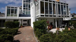 Ein modernes, zweistöckiges Gebäude mit großen Fenstern, einem Balkon und einem Ziegelweg, der zu einer Terrasse mit Gartenmöbeln führt.