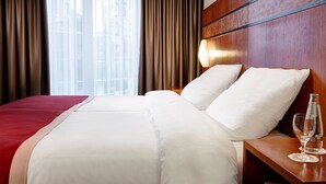 Ein Hotelzimmer mit einem großen Bett, einem hölzernen Kopfteil, einem Nachttisch mit einem Glas Wasser und Blick auf Gebäude durch transparente Vorhänge.
