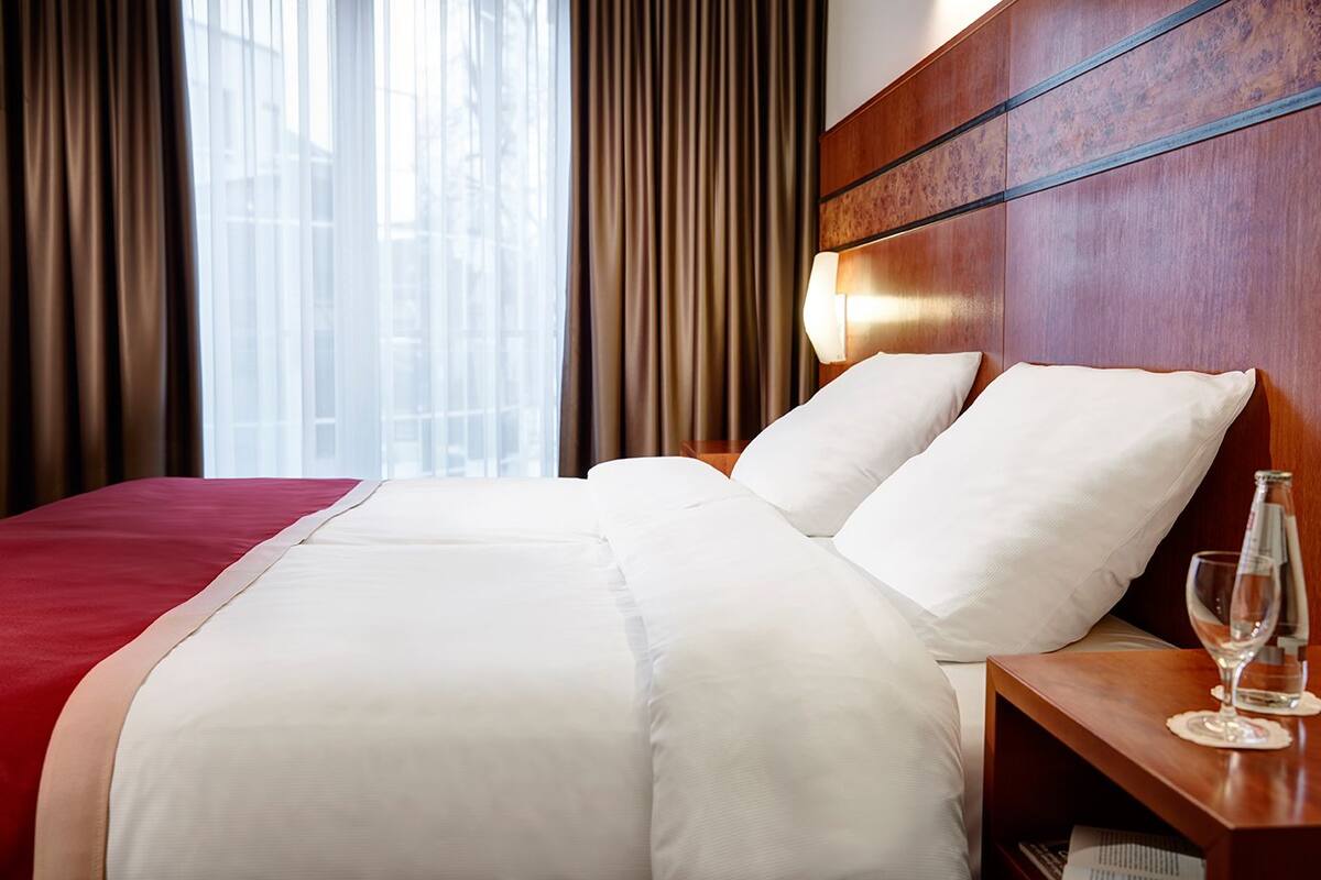 A hotel room with a large bed, a wooden headboard, a nightstand with a glass of water, and a view of buildings through sheer curtains.