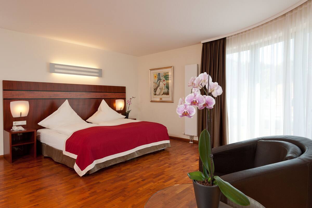 A hotel room with a wooden headboard, a bed with white bedding and a red accent, a bedside table with a lamp, a potted orchid, a large window with sheer curtains, and a leather armchair.