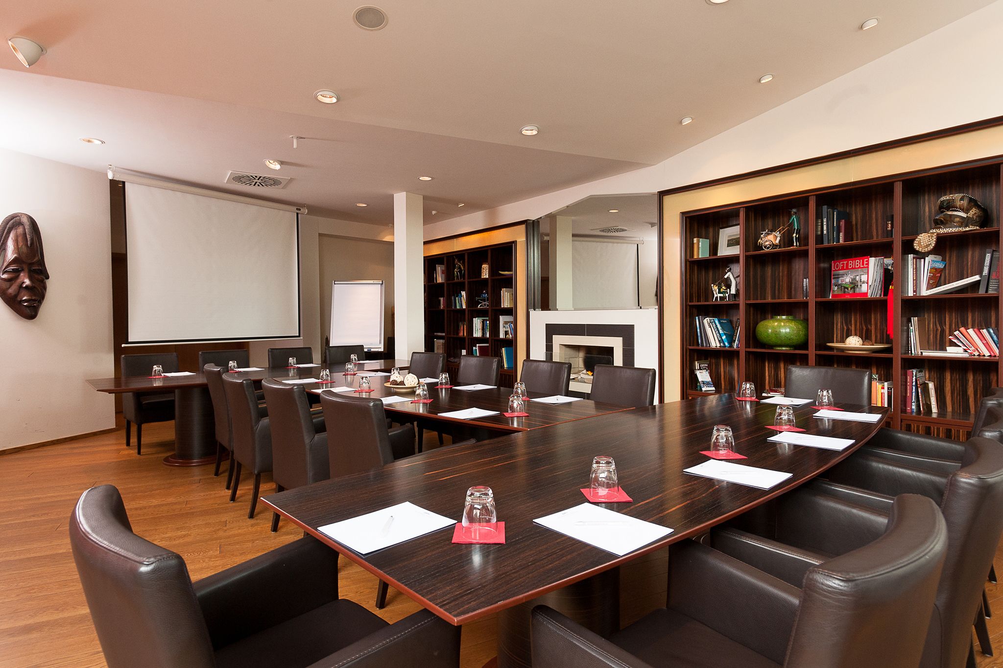 Une salle de conférence dotée d’une grande table en bois, de fauteuils en cuir, d’un écran de projection et d’une bibliothèque contenant divers ouvrages et objets décoratifs.