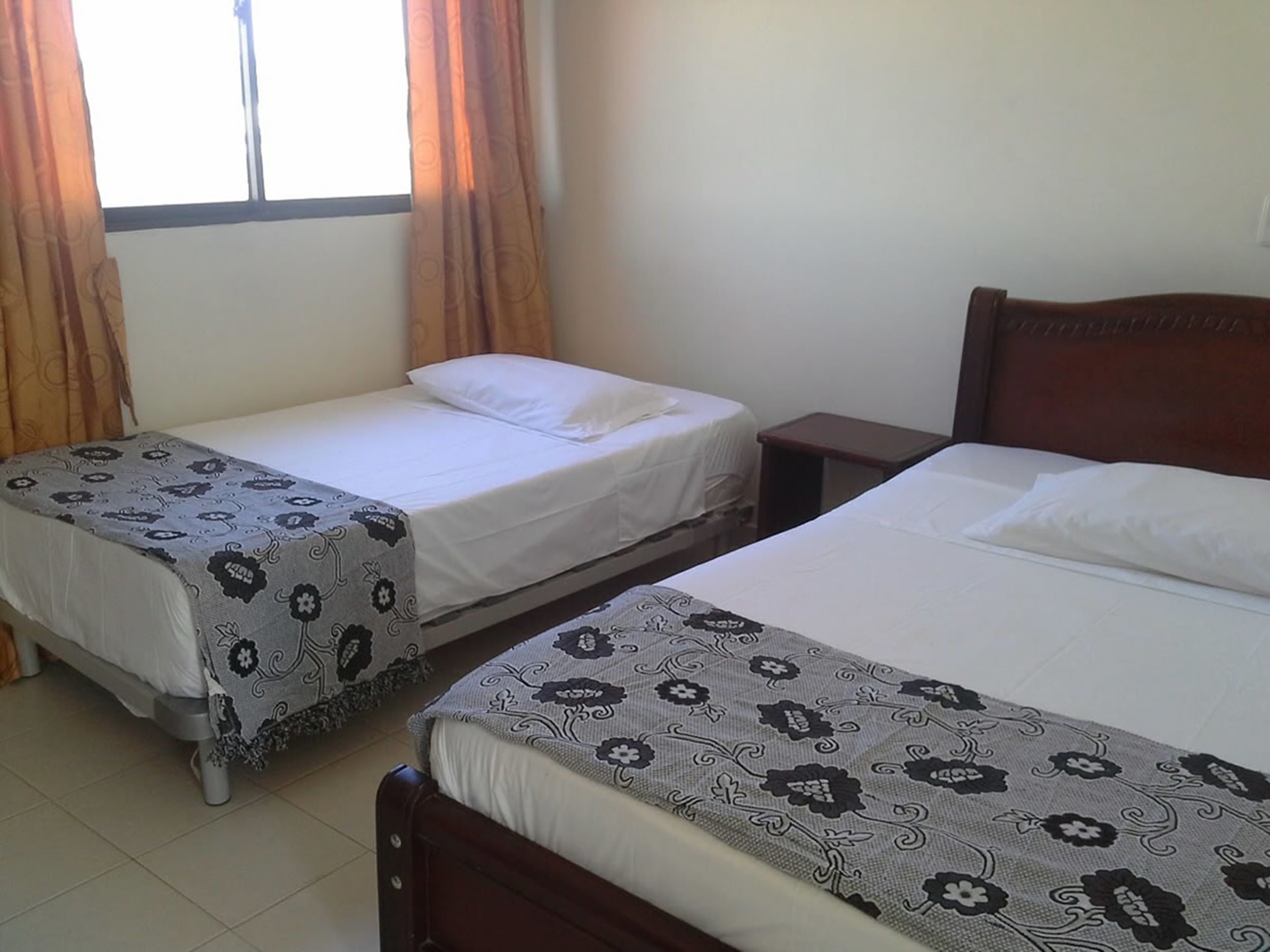 A room with two single beds, each with a patterned bedspread and white pillows. There is a small wooden nightstand between the beds, and a window with curtains on the left side.