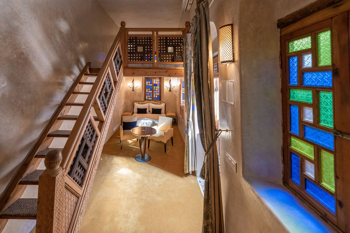 A narrow hallway with a wooden staircase, a dining area with a round table and chairs, and a window with colorful stained glass.