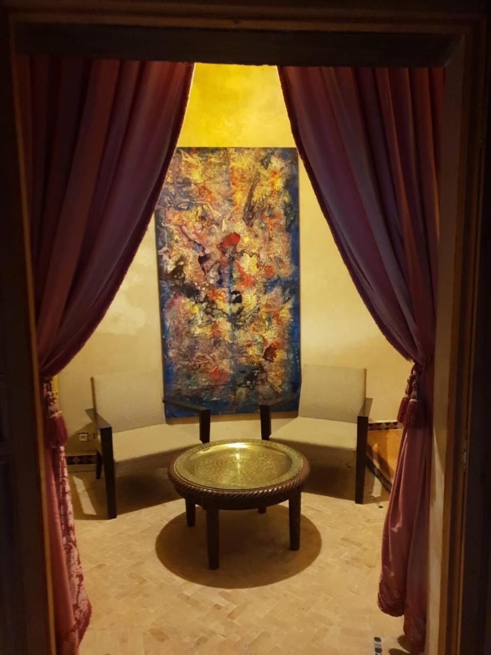 Quarto com uma grande pintura abstrata na parede, uma mesa redonda com tampo de vidro e cortinas roxas emoldurando a janela.