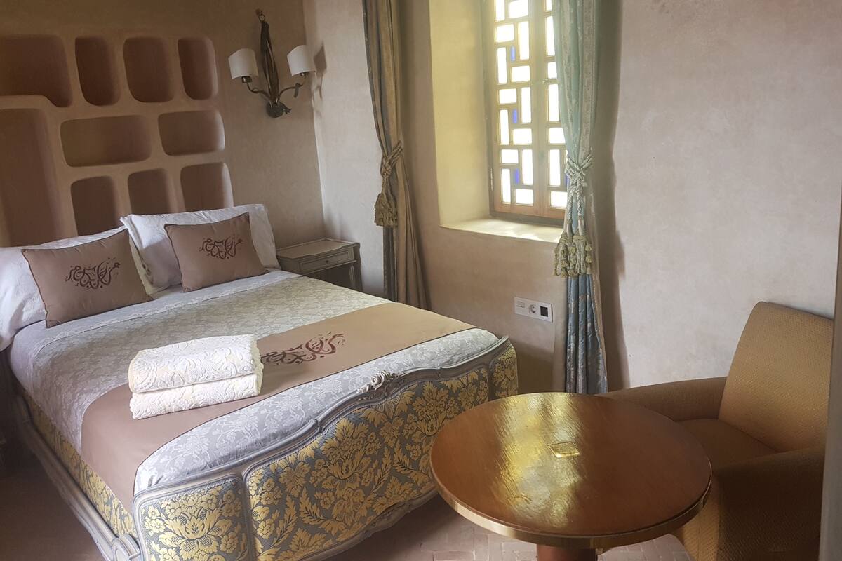 A bed with a headboard, pillows, a bedside table, a window with curtains, and a round table.