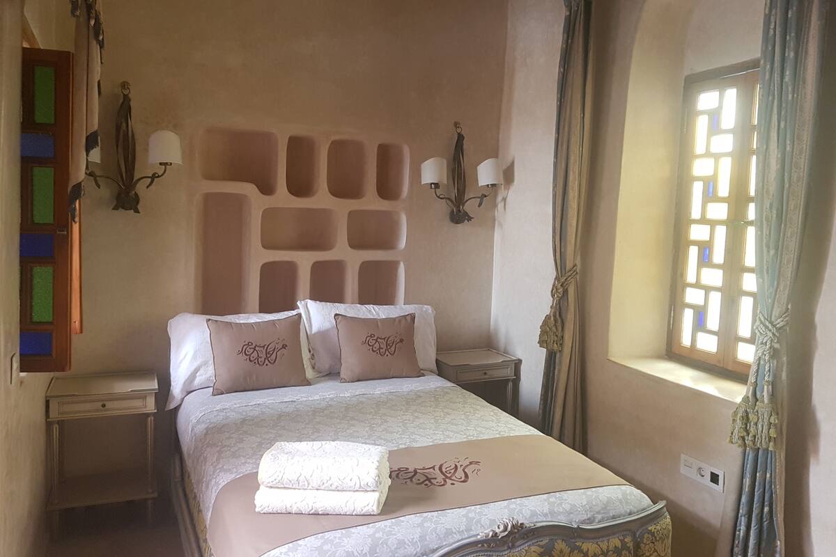 A bedroom with a large bed, two bedside tables, a chandelier, and a window with curtains.