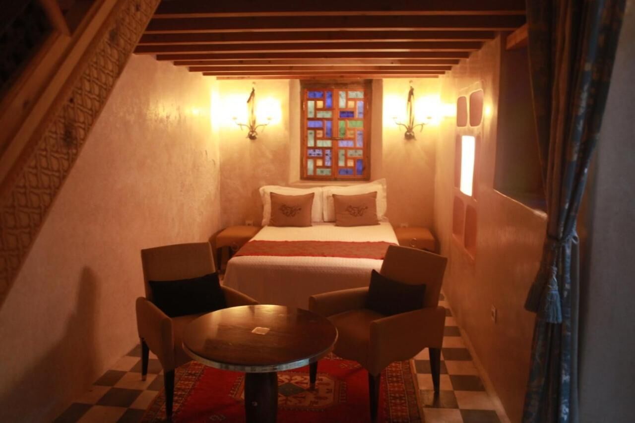 A cozy bedroom with a bed, two chairs, a small table, and a decorative window.