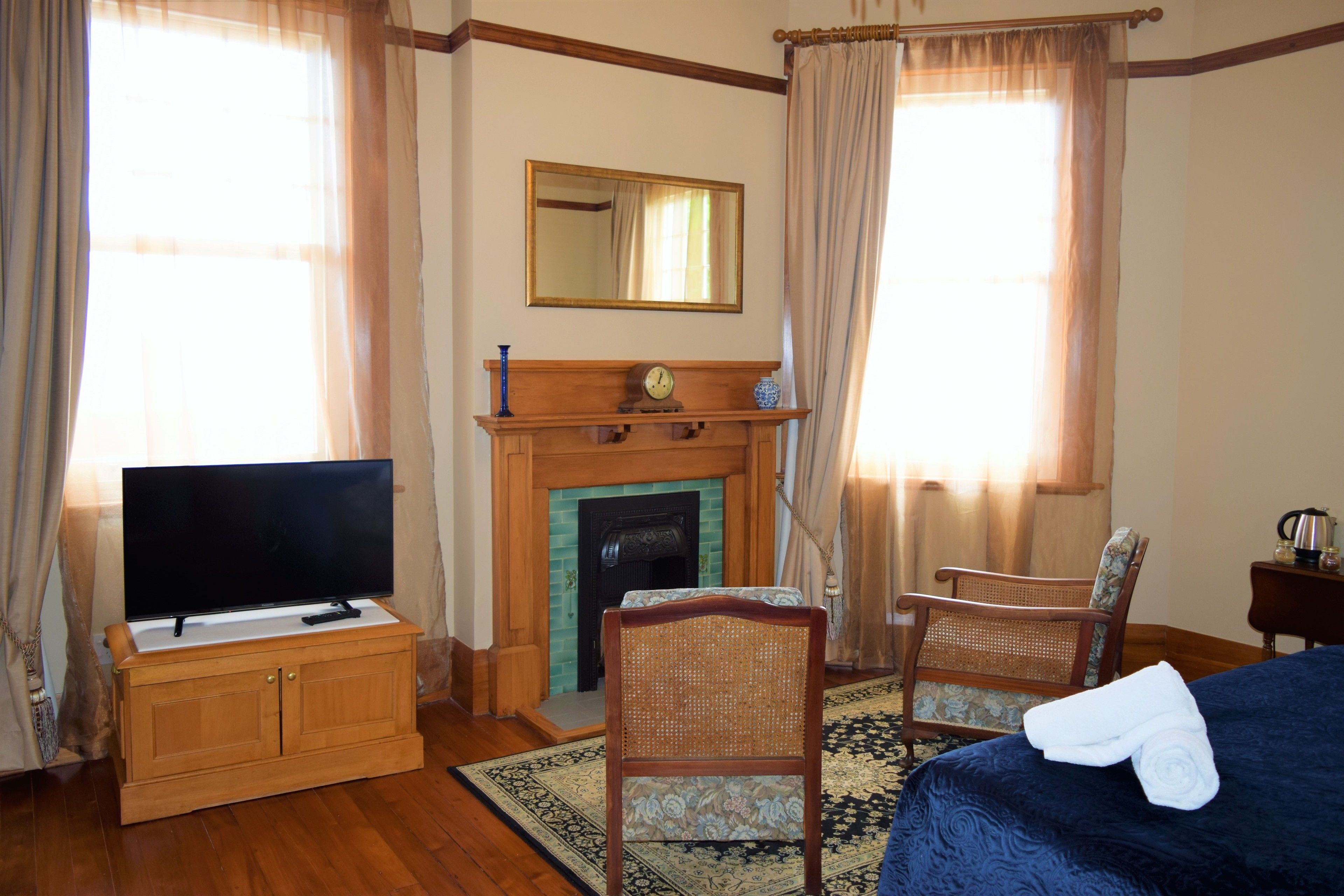 A hotel room with a television on a wooden cabinet, a fireplace, a bed with a blue bedspread, a chair, and a small table with a kettle.
