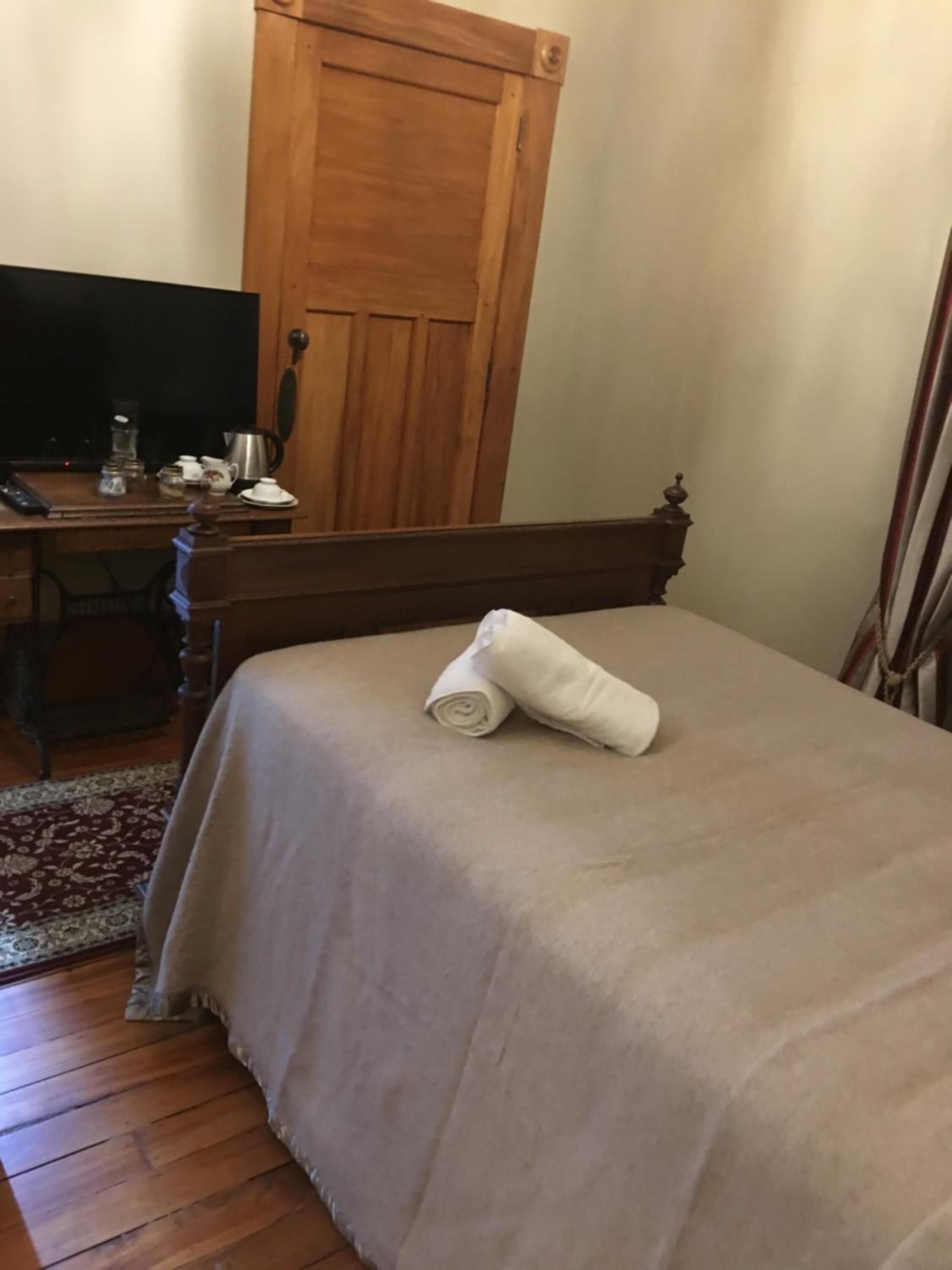 Un lit bien fait avec une serviette pliée, un bureau en bois avec un ordinateur et une cafetière, et une porte en bois dans une pièce au plancher en bois.