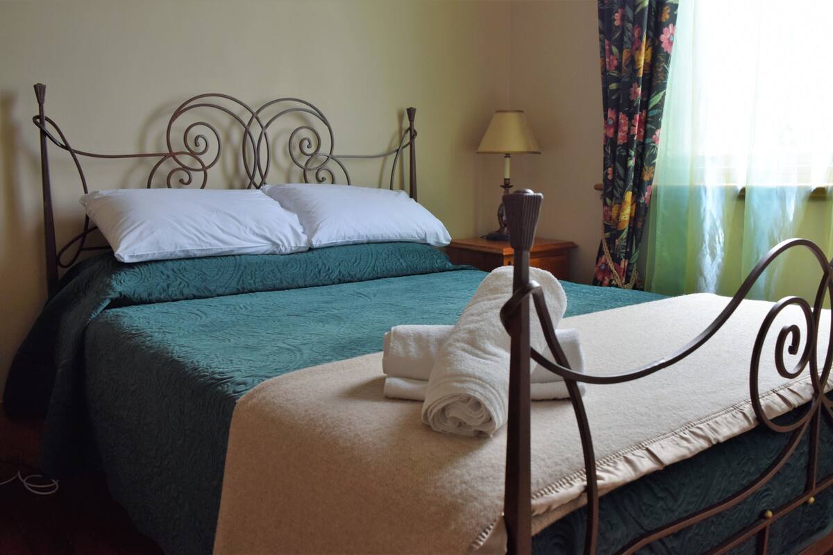 A neatly made bed with a teal bedspread, white pillows, and a folded white towel on the side.