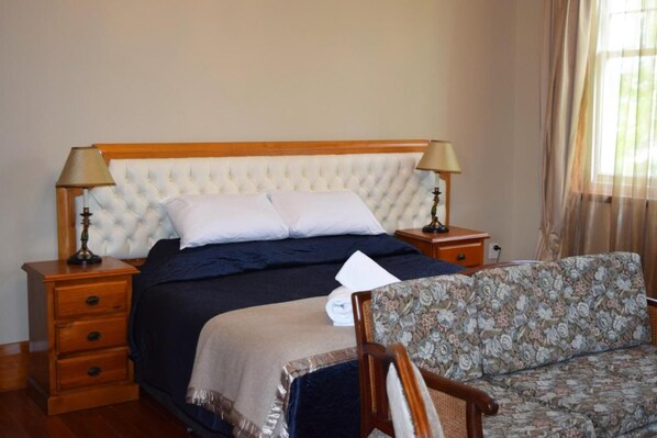 Una habitación de hotel con una cama grande, dos mesitas de noche con lámparas, una mesita de noche de madera, un sofá floral y una ventana con cortinas.