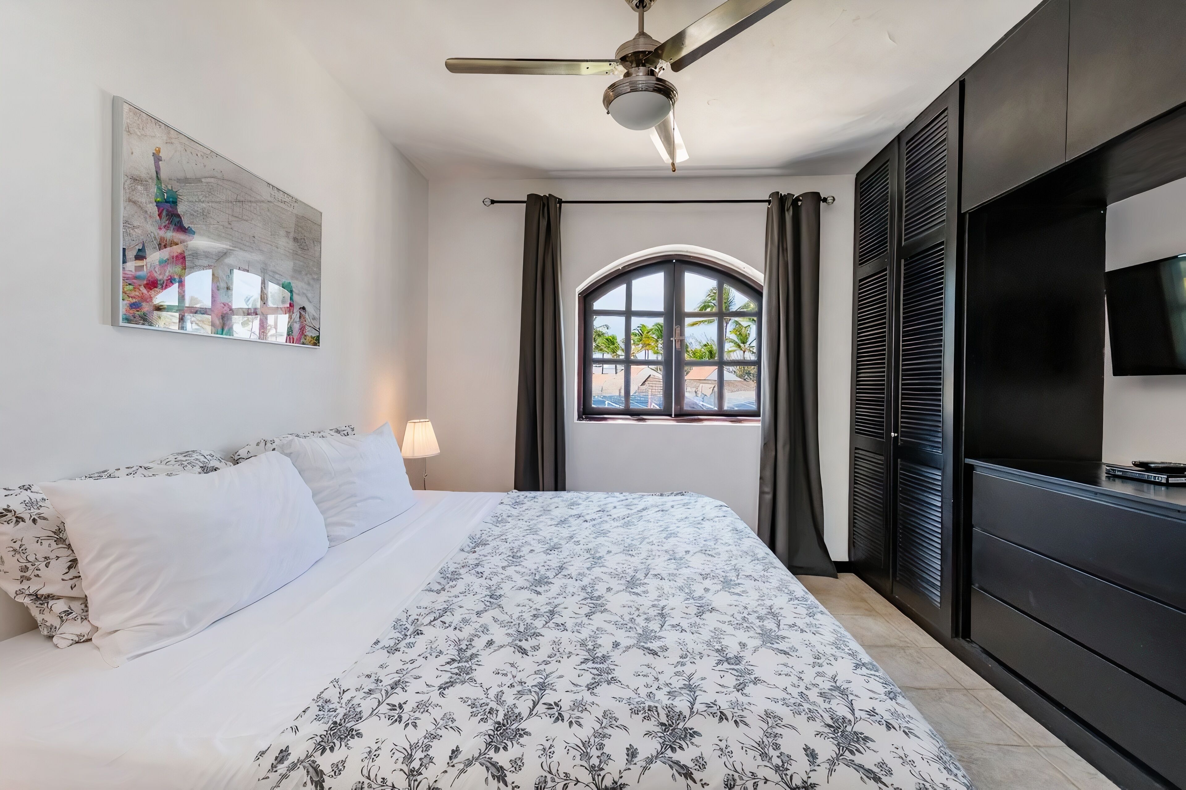 Ein Schlafzimmer mit Bett, Deckenventilator, Fernseher, Fenster mit Vorhängen und einem Bild an der Wand.