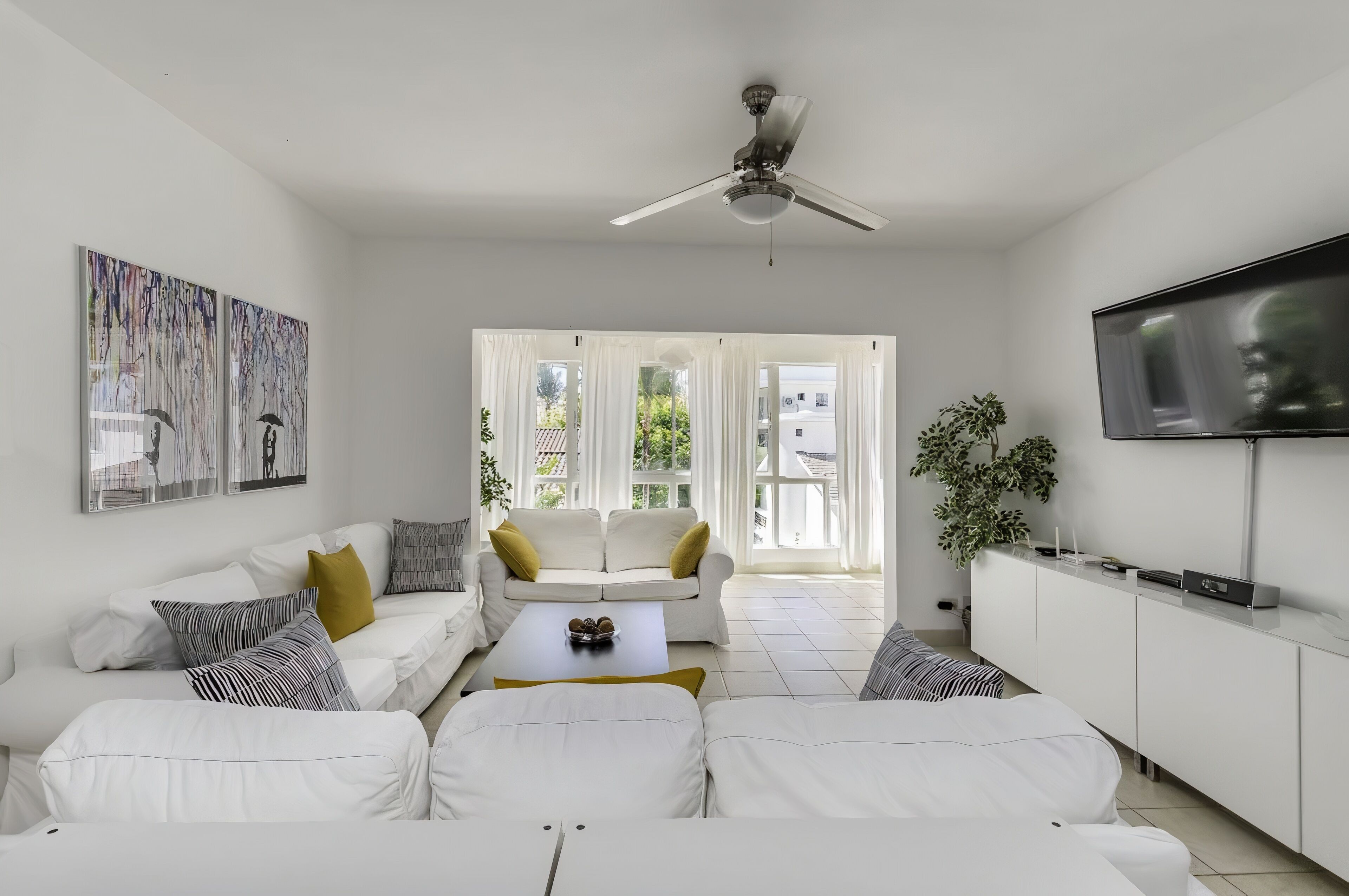 A modern living room with white furniture, a ceiling fan, a flat-screen TV, and artwork on the walls.