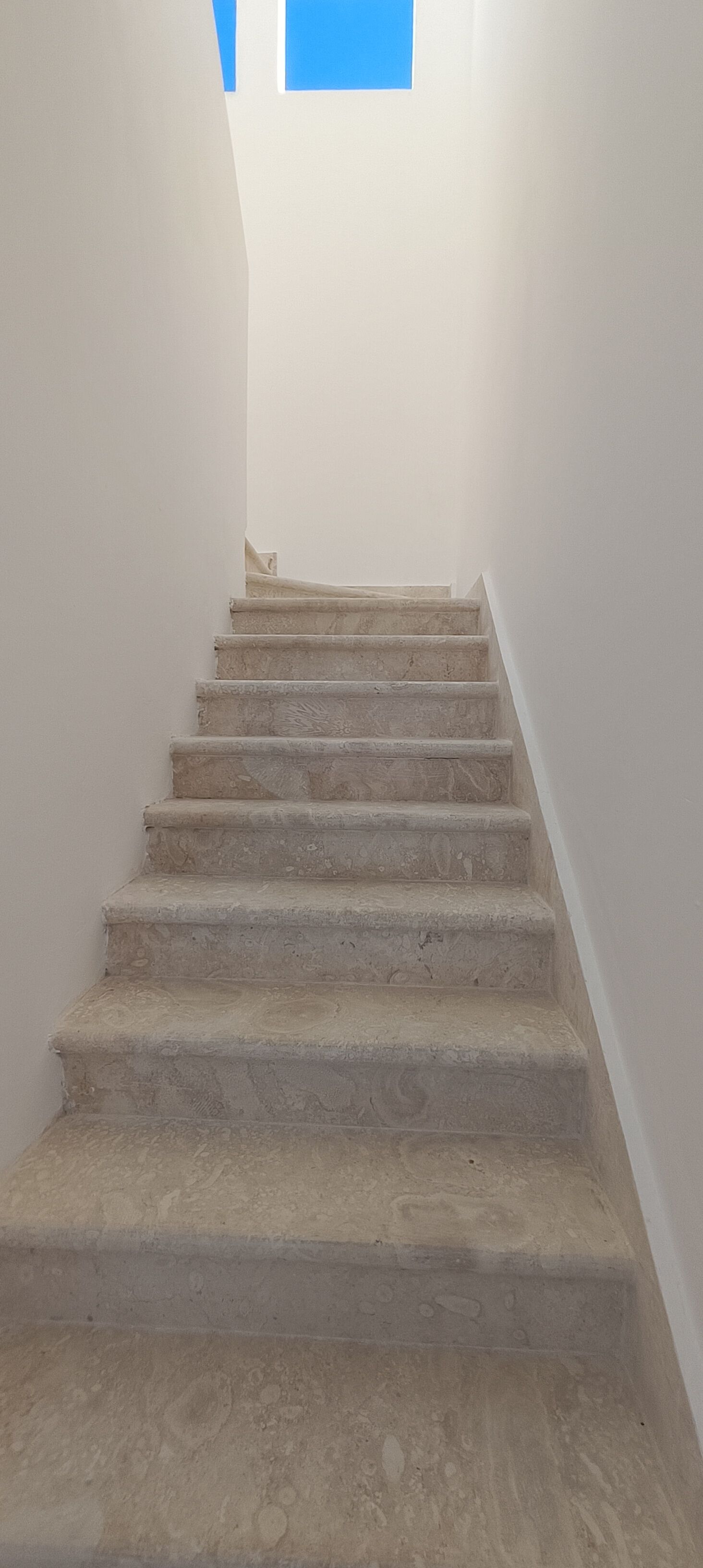 A staircase with a plain white wall on one side and a blue sky visible through a window at the top.