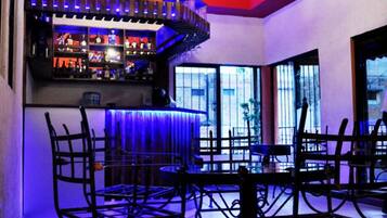 Un área de bar con techo alto, vigas a la vista y una lámpara de araña. Hay varios taburetes de bar y una barra con estantes llenos de botellas.