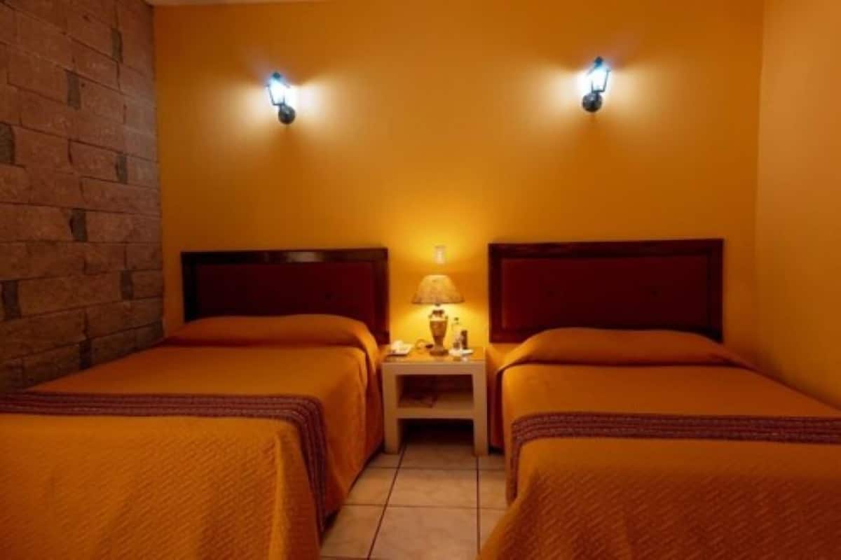 A hotel room with two beds, a nightstand with a lamp, and two wall-mounted light fixtures.