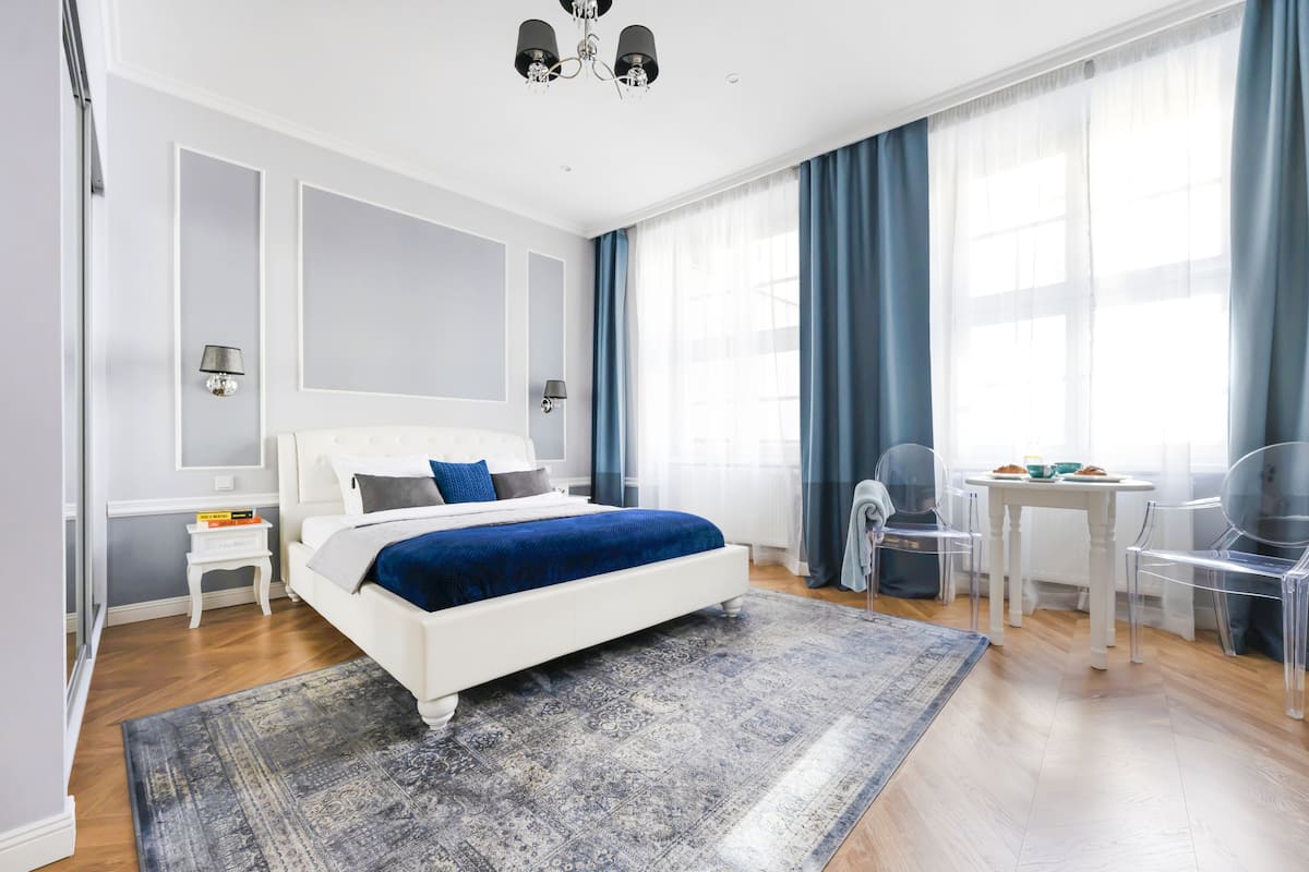 Un dormitorio moderno con cama blanca, ropa de cama azul, una mesita de noche pequeña, un escritorio con sillas y un ventanal con cortinas.