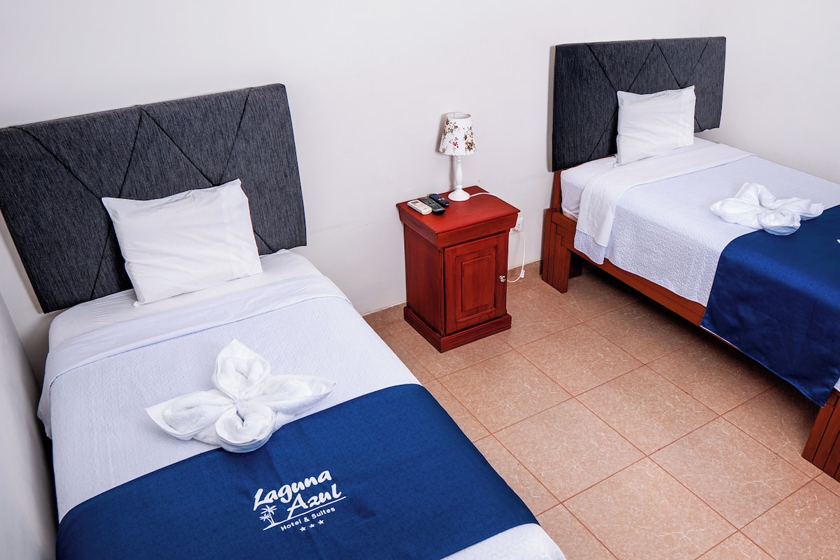 Dos camas con cabeceras grises, almohadas blancas y sábanas azules con un logo. Hay una mesita de noche de madera con una lámpara y un control remoto entre las camas.