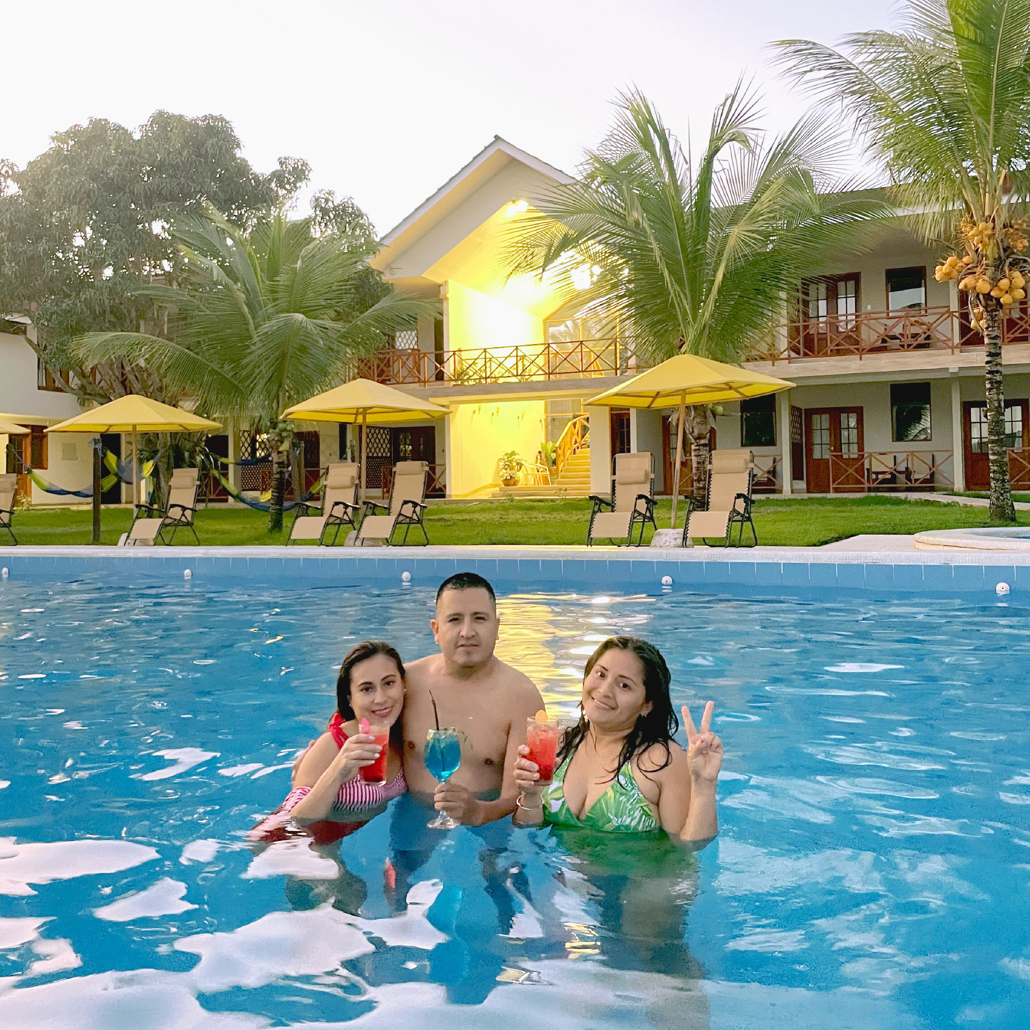 A swimming pool with lounge chairs and umbrellas, a two-story building with balconies, and palm trees in the background.