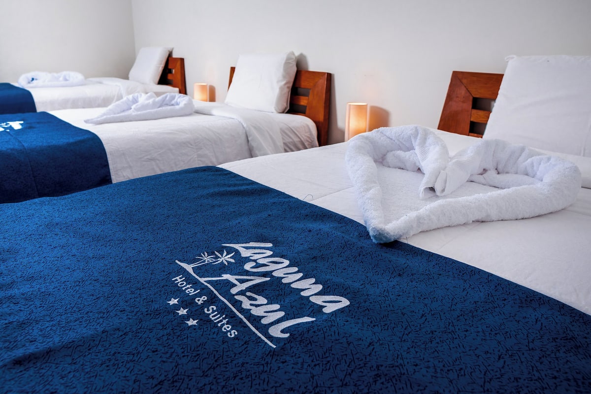 Habitación de hotel con dos camas, cada una con ropa blanca y cabeceros de madera. Una cama tiene una manta azul con el nombre y el logo del hotel.