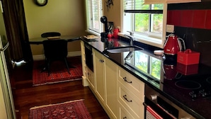 A kitchen with white cabinets, a black countertop, and a red kettle. There is a window with blinds, a clock on the wall, and a rug on the floor. - Matai Huka Boutique Accommodation (Paraparaumu)