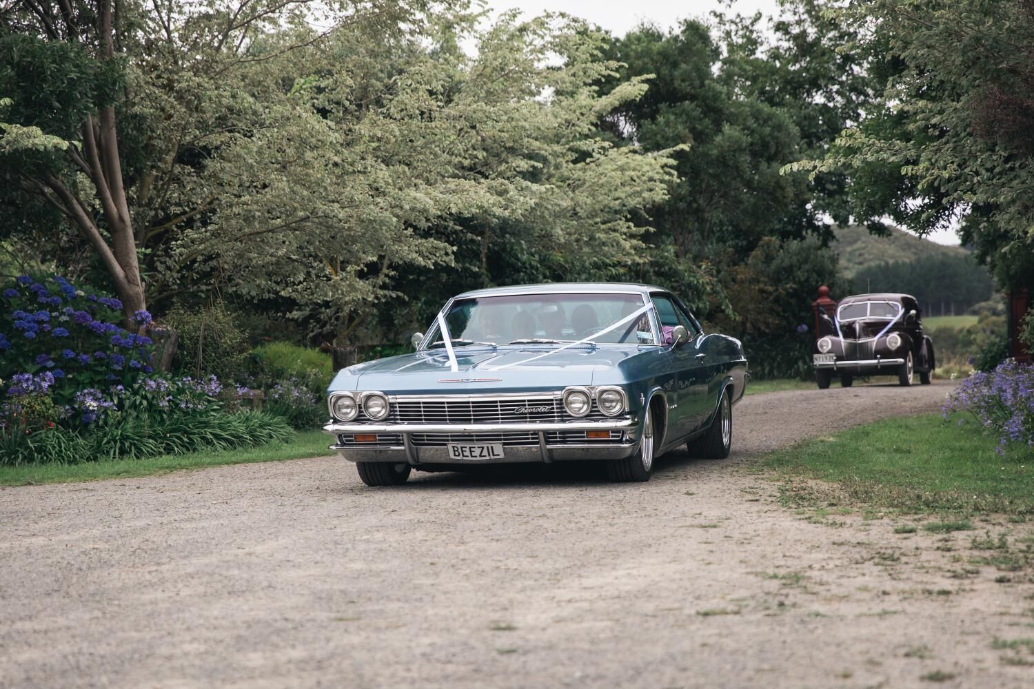 Une voiture ancienne sur une route de campagne, entourée d’une végétation luxuriante et de fleurs.