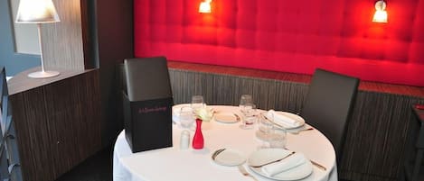 Un comedor con pared tapizada en rojo, una mesa blanca dispuesta con platos, vasos y cubiertos, y un jarrón rojo con flores blancas.