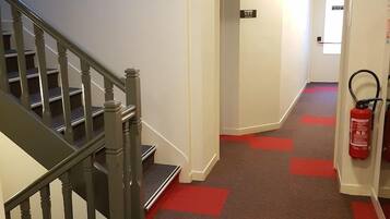 Un couloir d’hôtel avec un escalier, un extincteur et un panneau indiquant la direction des chambres.