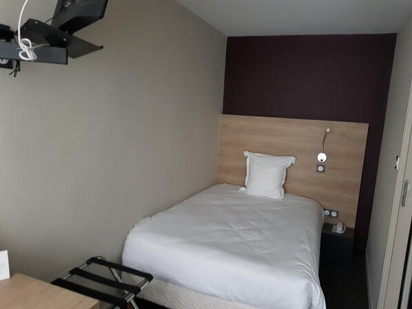 Une petite chambre d’hôtel avec un lit simple, un bureau et un luminaire fixé au mur.