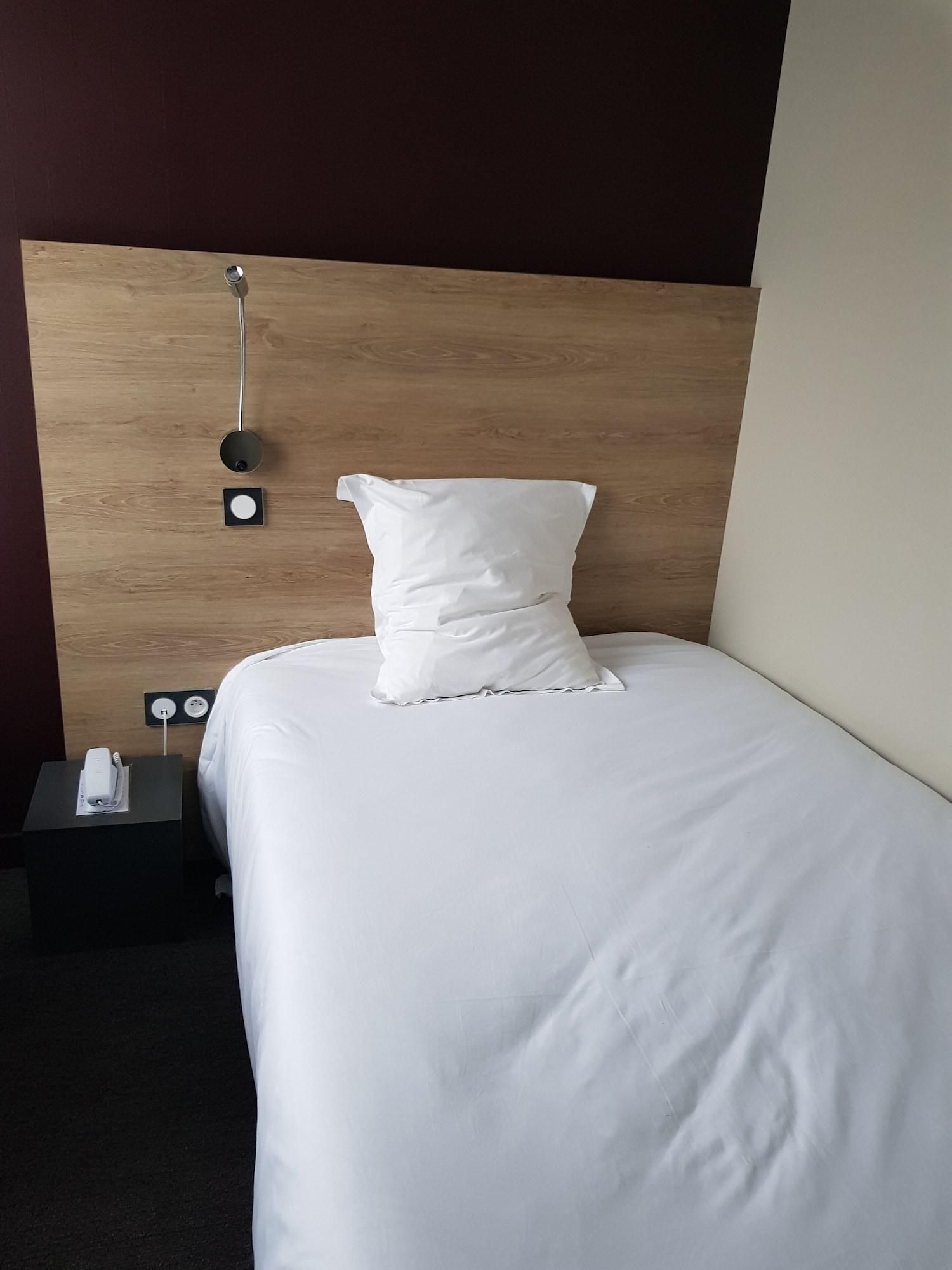 Una cama bien tendida con ropa blanca y almohadas, cabecera de madera y mesita de noche con un control remoto.