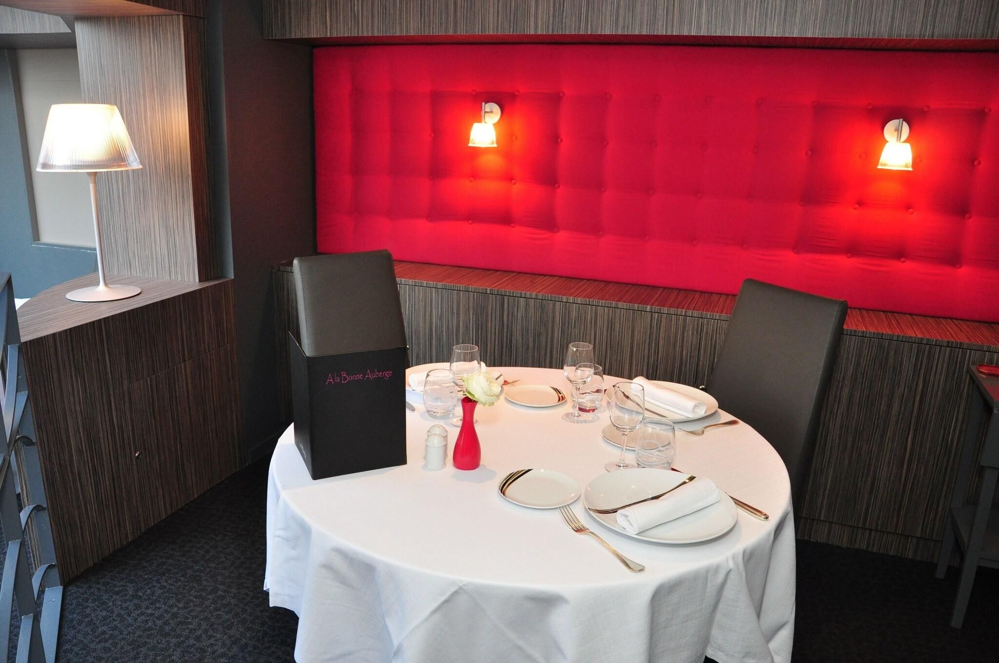 Mesa vestida con mantel blanco, platos, vasos y cubiertos. Se ve una pared tapizada de rojo y una lámpara al fondo.