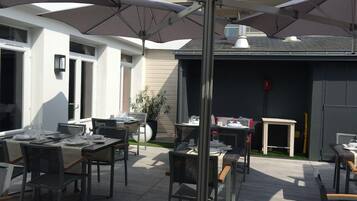 Un espace extérieur aménagé pour les repas, avec des tables, des chaises, des parasols et un bâtiment en arrière-plan.