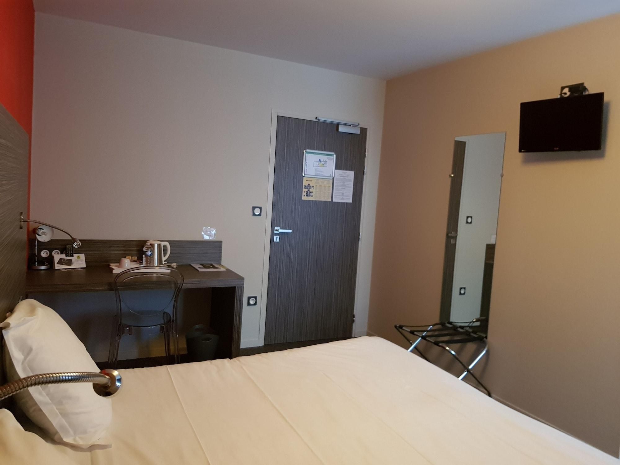A hotel room with a bed, a desk, a chair, a mirror, and a door.