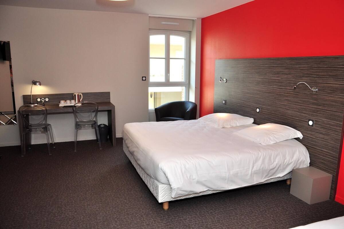A hotel room with a bed, a desk, a chair, and a red accent wall.
