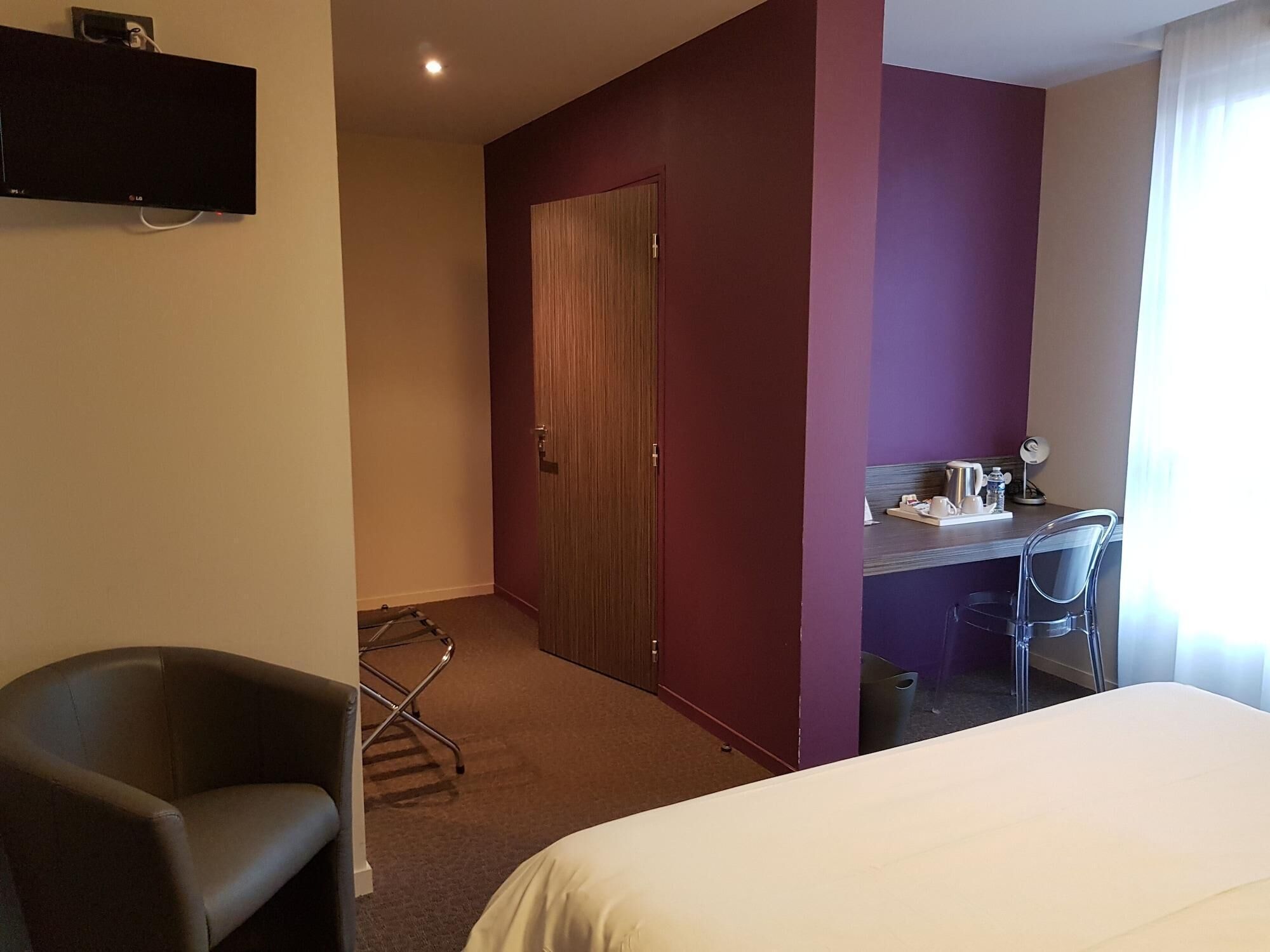 Een hotelkamer met een bed, een bureau met stoel, een aan de muur bevestigde televisie en een deur naar een andere kamer.