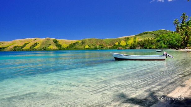 Een boot op een helder turquoise strand, met op de achtergrond groene heuvels en palmbomen.