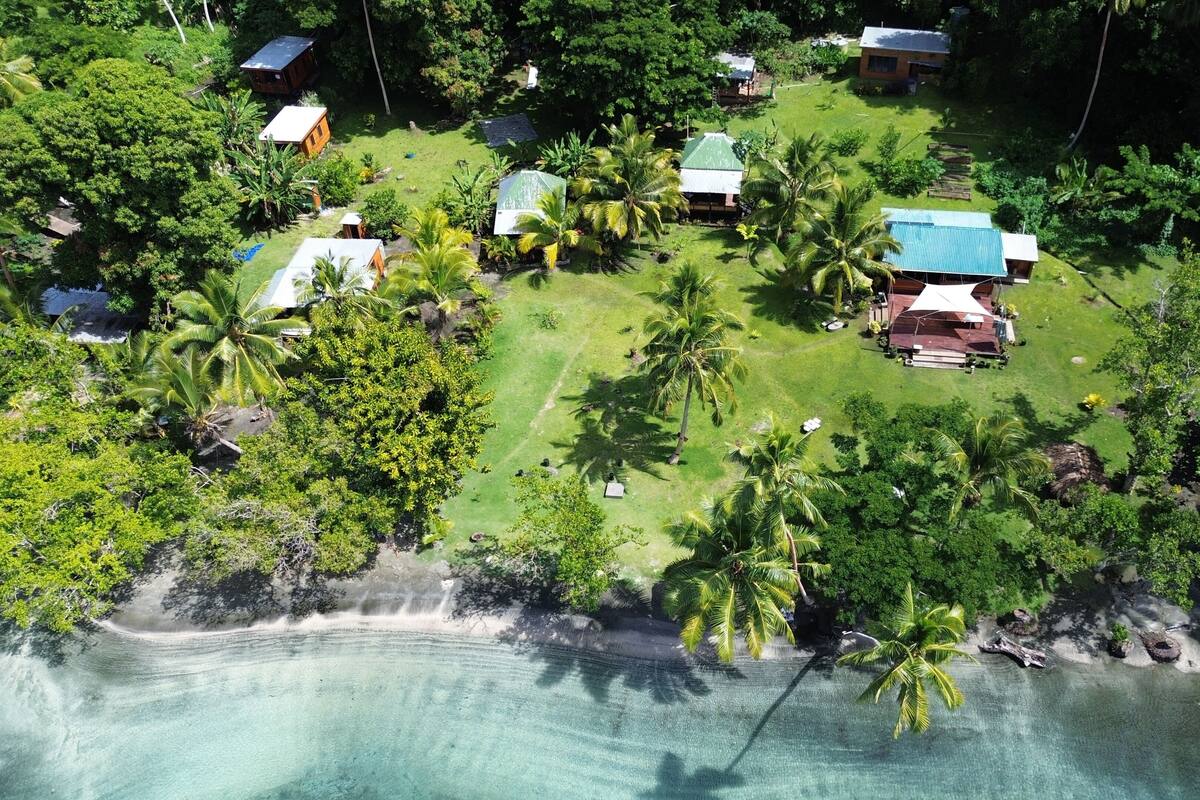 Vue aérienne d’un complexe hôtelier tropical doté de plusieurs bâtiments de style bungalow, d’une végétation luxuriante et d’une plage de sable fin aux eaux turquoise.