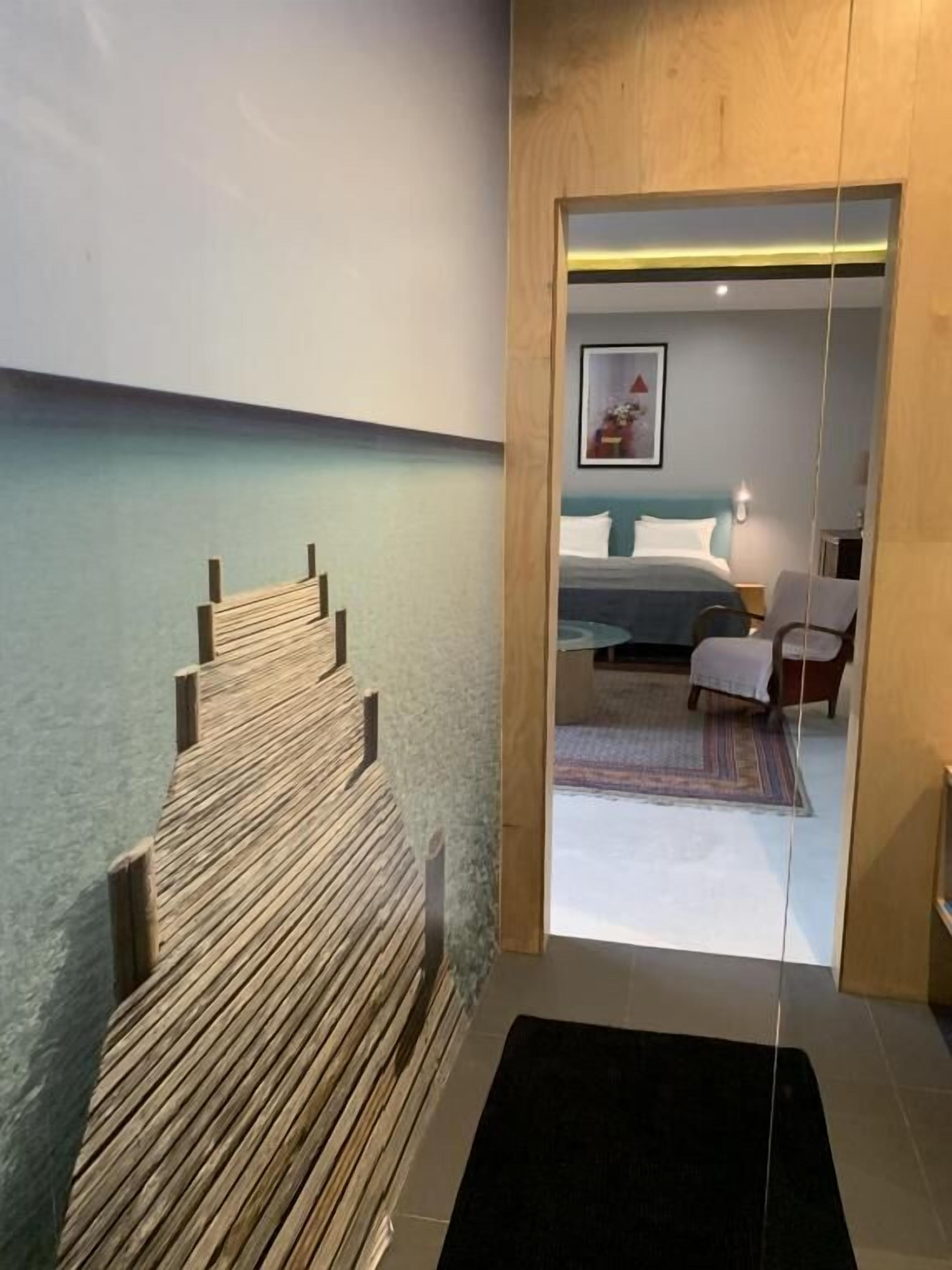 A modern hotel room with a wooden wall feature, a bed with white bedding, a small seating area, and a visible bathroom.