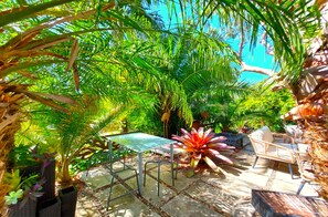 A tropical garden patio with a metal table and chairs, surrounded by lush greenery and a large decorative plant. - Luxury Escape (Palm Beach)