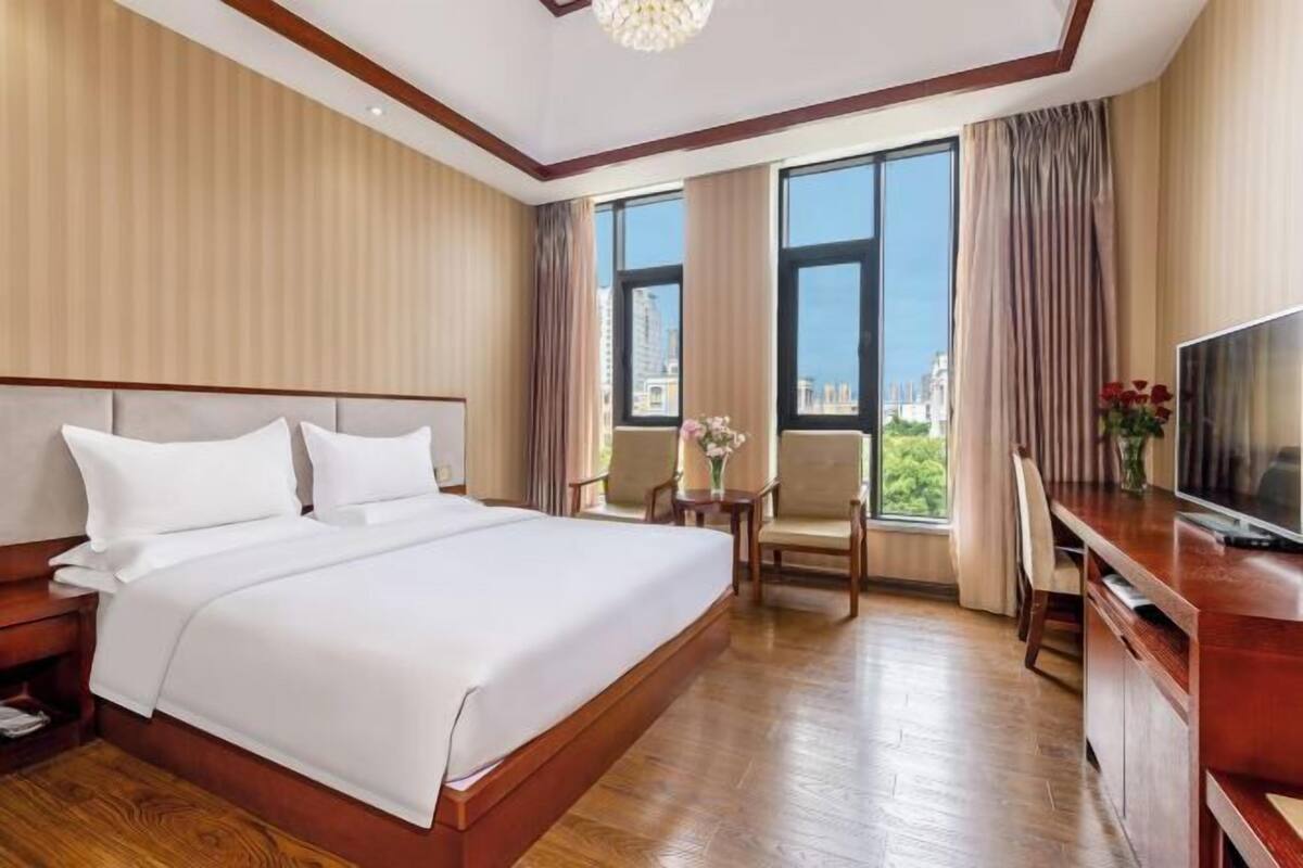 Une chambre d’hôtel avec un grand lit, un bureau avec une télévision et une vue sur la ville grâce à de grandes fenêtres.