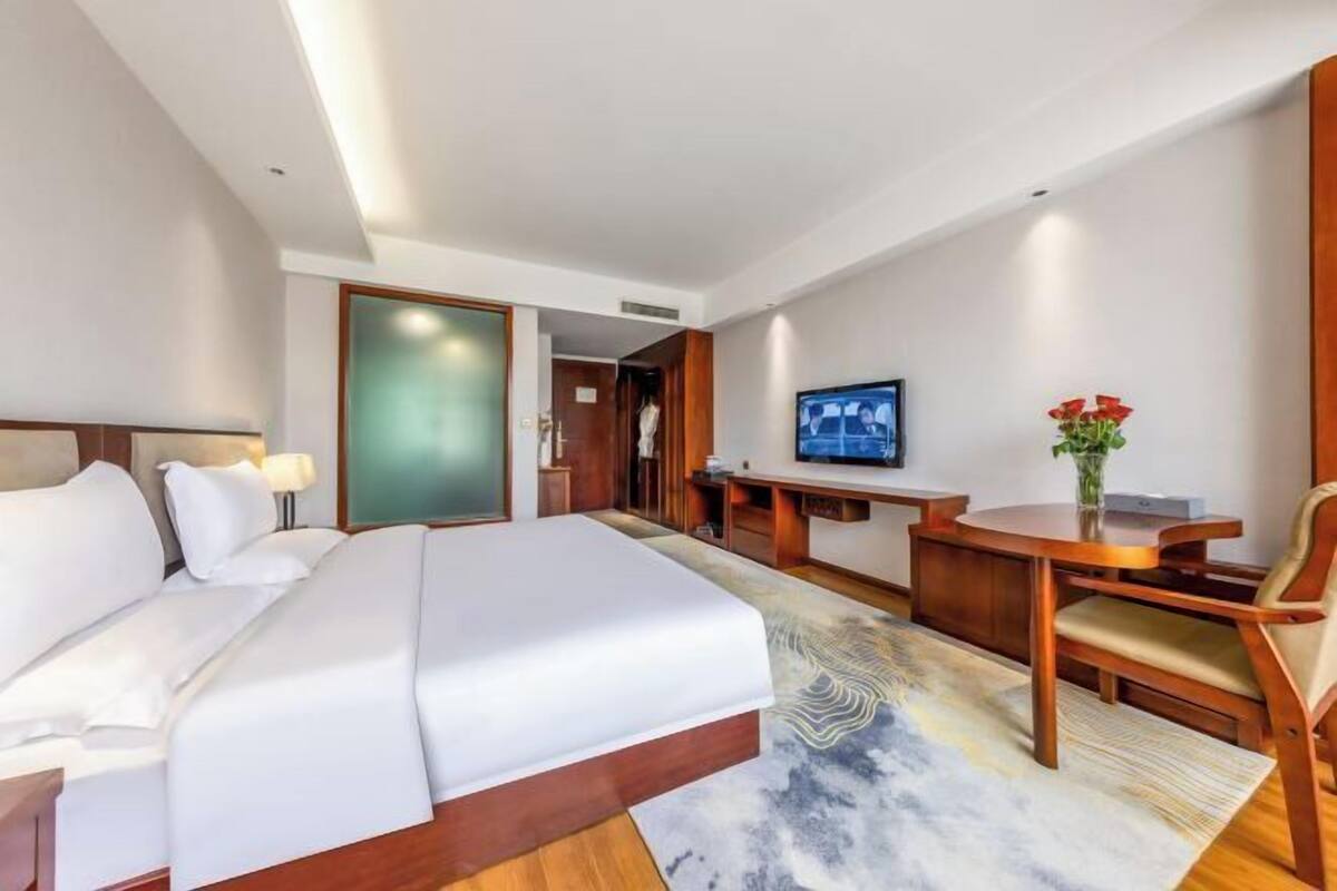 Une chambre d’hôtel avec un grand lit, un bureau avec une chaise, une télévision et une petite table sur laquelle est posé un vase de fleurs.