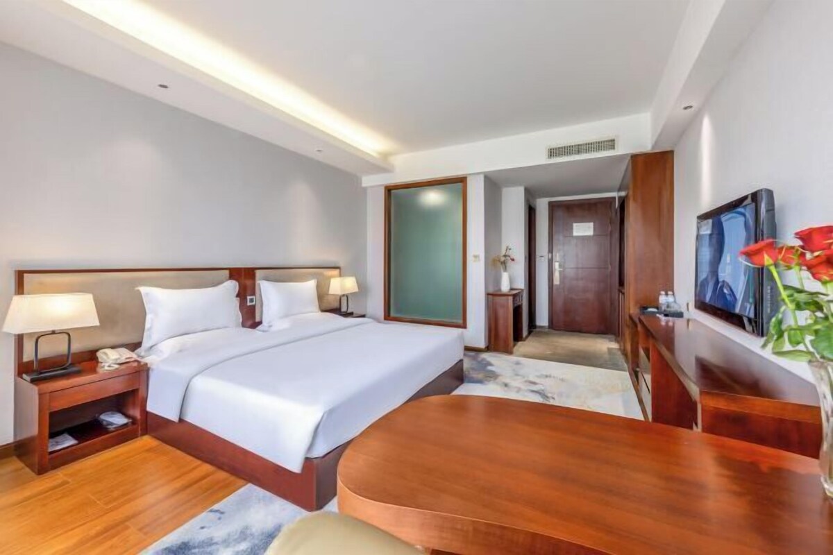 Une chambre d’hôtel avec un grand lit, des tables de chevet avec des lampes, un bureau en bois, une télévision et un vase rempli de fleurs.