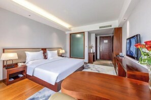 Une chambre d’hôtel avec un grand lit, des tables de chevet avec des lampes, un bureau en bois, une télévision et un vase rempli de fleurs.