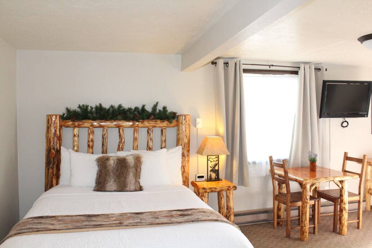 Et hotelværelse med en rustik træhovedgavl, en seng med hvide sengetøj, et lille natbord med en lampe, et træspisebord med stole, og et tv ophængt på væggen.