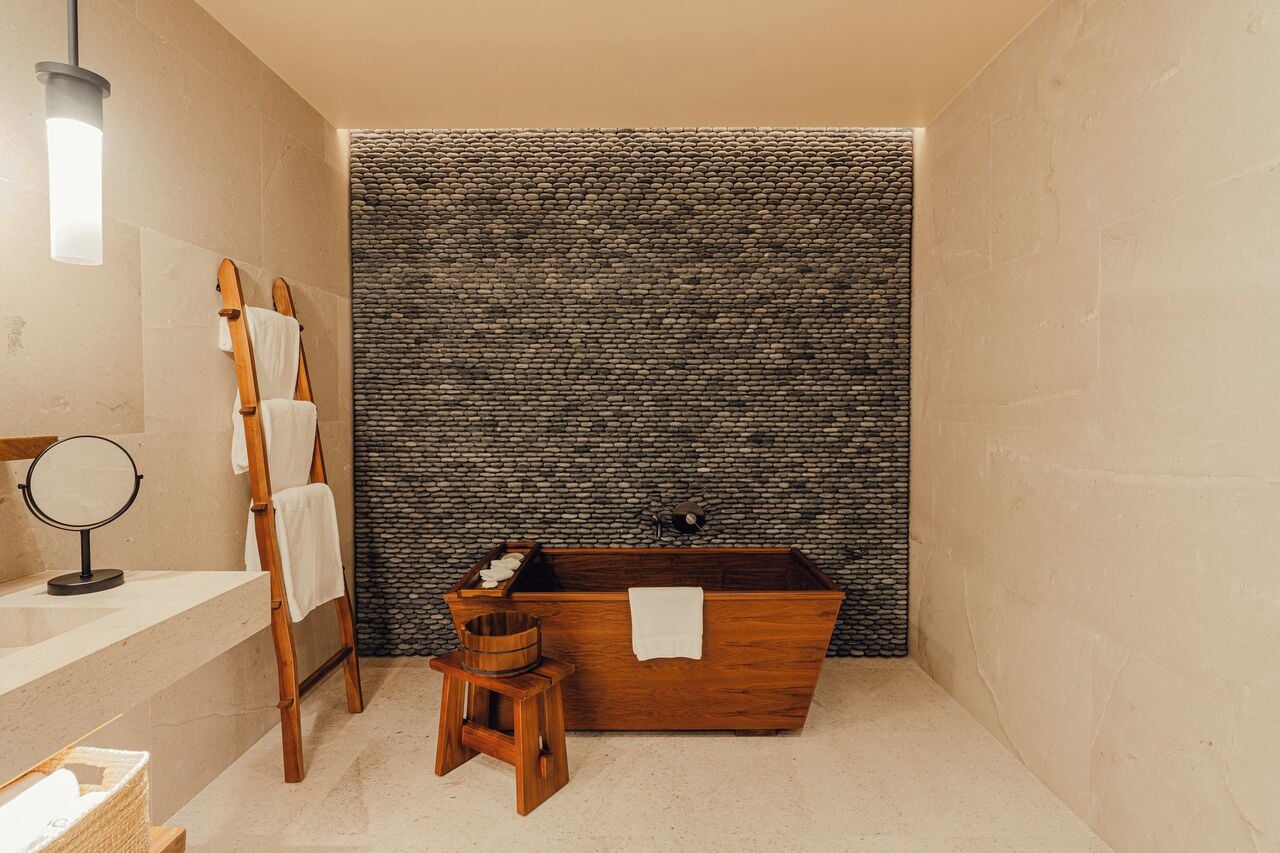 A bathroom with a stone wall, a wooden bathtub, a wooden stool, and a wooden ladder.