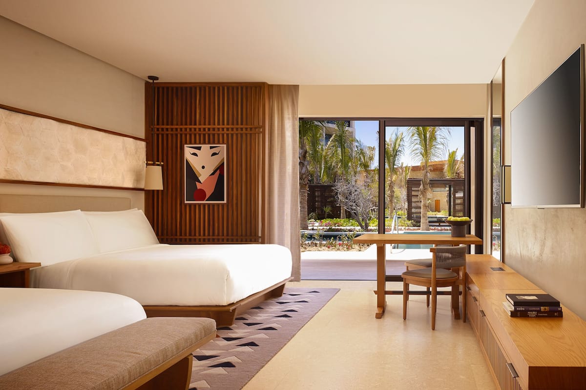 A modern hotel room with two beds, a dining area, and a view of a pool and palm trees.