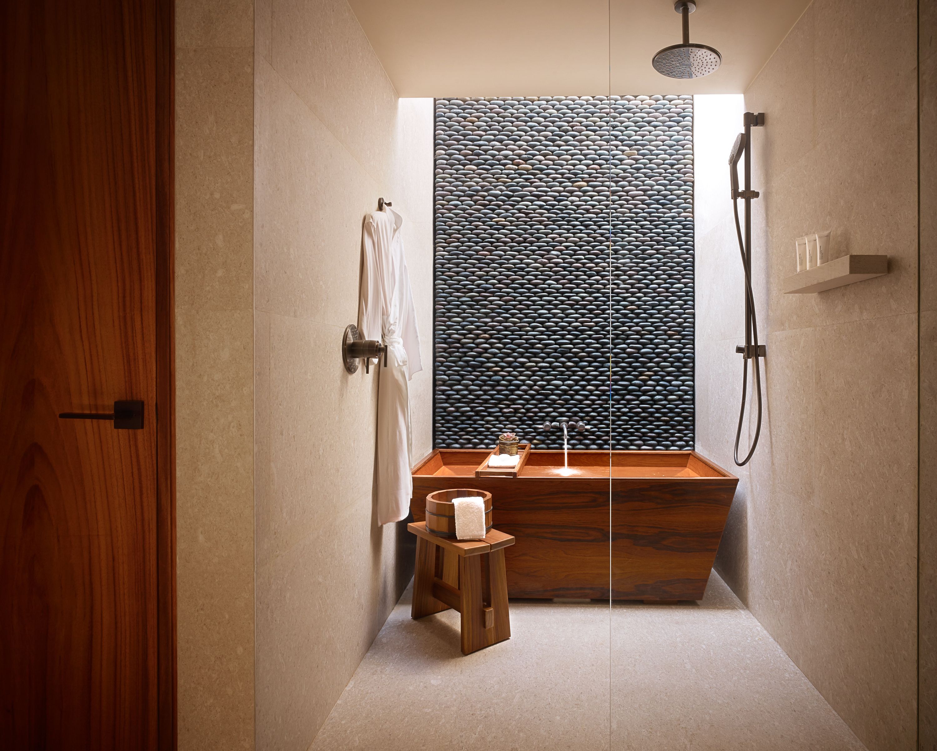 Un bagno moderno con una vasca da bagno in legno, un piccolo sgabello di legno, un porta asciugamani e una doccia con parete a parete in pietra.